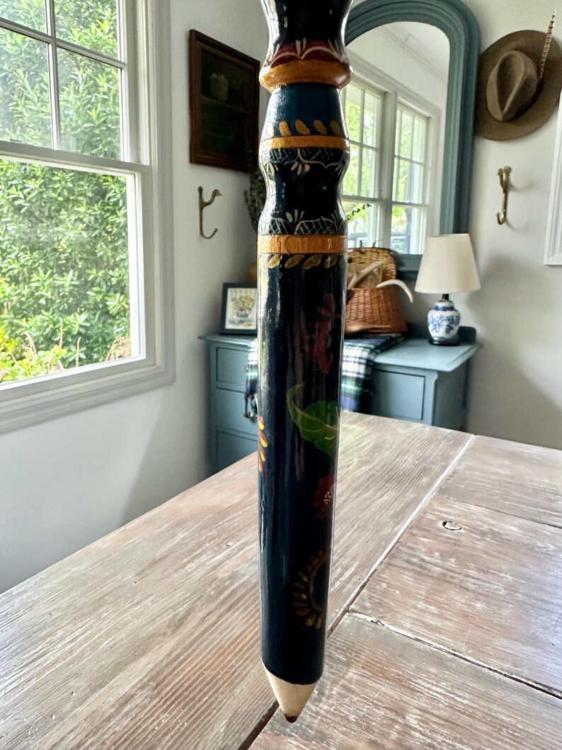 May include: A tall, decorative wooden pencil with a painted design. The pencil is primarily dark blue with colorful floral and geometric patterns. The tip is a light beige color, and the pencil stands upright on a wooden table.