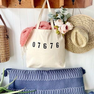 May include: A white canvas tote bag with black numbers "07676" printed on the front. The bag is hanging on a wooden shelf with a blue and white patterned pillow in the foreground.