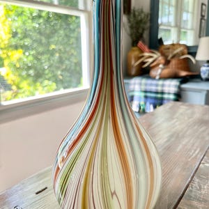 K&ouml;nnte beinhalten: Eine dekorative Glasvase mit bauchigem K&ouml;rper und schlankem Hals. Die Vase hat vertikale Streifen in Blau-, Orange-, Gr&uuml;n- und Braunt&ouml;nen auf cremefarbenem Hintergrund. Die Vase steht auf einer verwitterten Holzoberfl&auml;che.