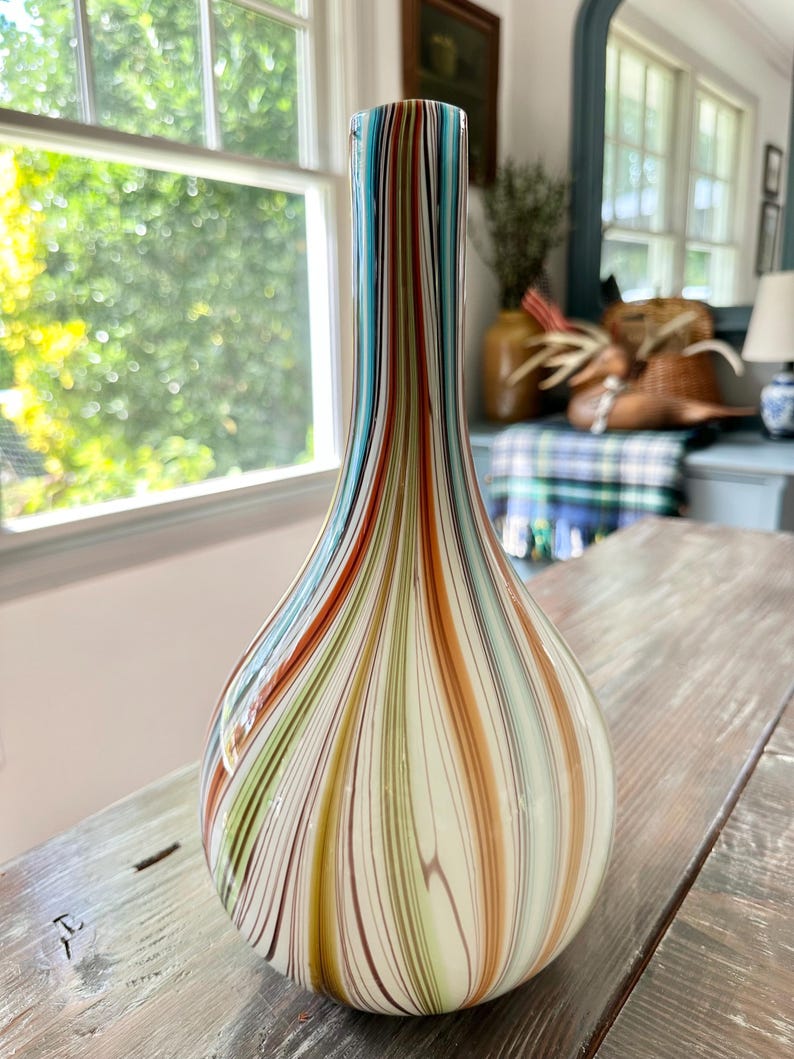 K&ouml;nnte beinhalten: Eine dekorative Glasvase mit bauchigem K&ouml;rper und schlankem Hals. Die Vase hat vertikale Streifen in Blau-, Orange-, Gr&uuml;n- und Braunt&ouml;nen auf cremefarbenem Hintergrund. Die Vase steht auf einer verwitterten Holzoberfl&auml;che.