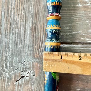 May include: A hand-painted wooden object with a blue base and colorful floral and geometric designs. The object is measured with a wooden ruler, showing a length of approximately 2 inches.
