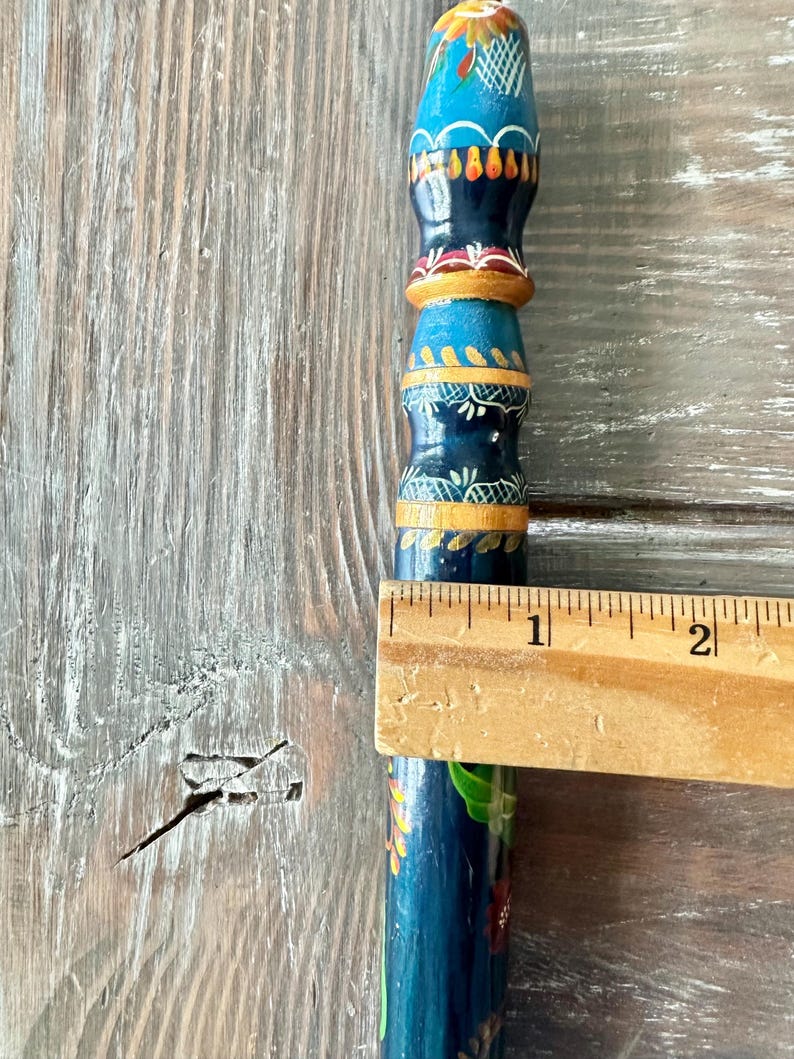 May include: A hand-painted wooden object with a blue base and colorful floral and geometric designs. The object is measured with a wooden ruler, showing a length of approximately 2 inches.