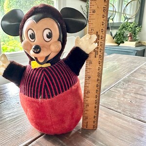 May include: Vintage Mickey Mouse plush toy. The toy has a brown head and ears, a red and black striped torso, and a red bottom. It has a yellow bow tie and outstretched arms. The ruler next to it measures approximately 8 inches.