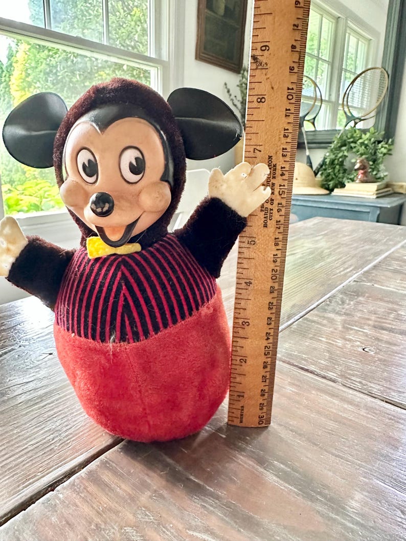 May include: Vintage Mickey Mouse plush toy. The toy has a brown head and ears, a red and black striped torso, and a red bottom. It has a yellow bow tie and outstretched arms. The ruler next to it measures approximately 8 inches.