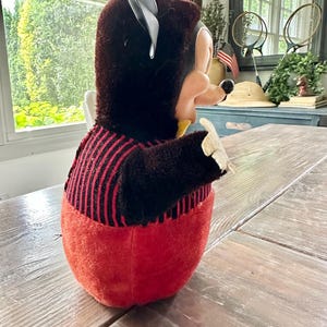 May include: A vintage plush toy in the shape of a cartoon character, featuring a black head with large ears and a red body. The toy has a black and red striped shirt and a yellow collar. The toy is sitting on a wooden table.