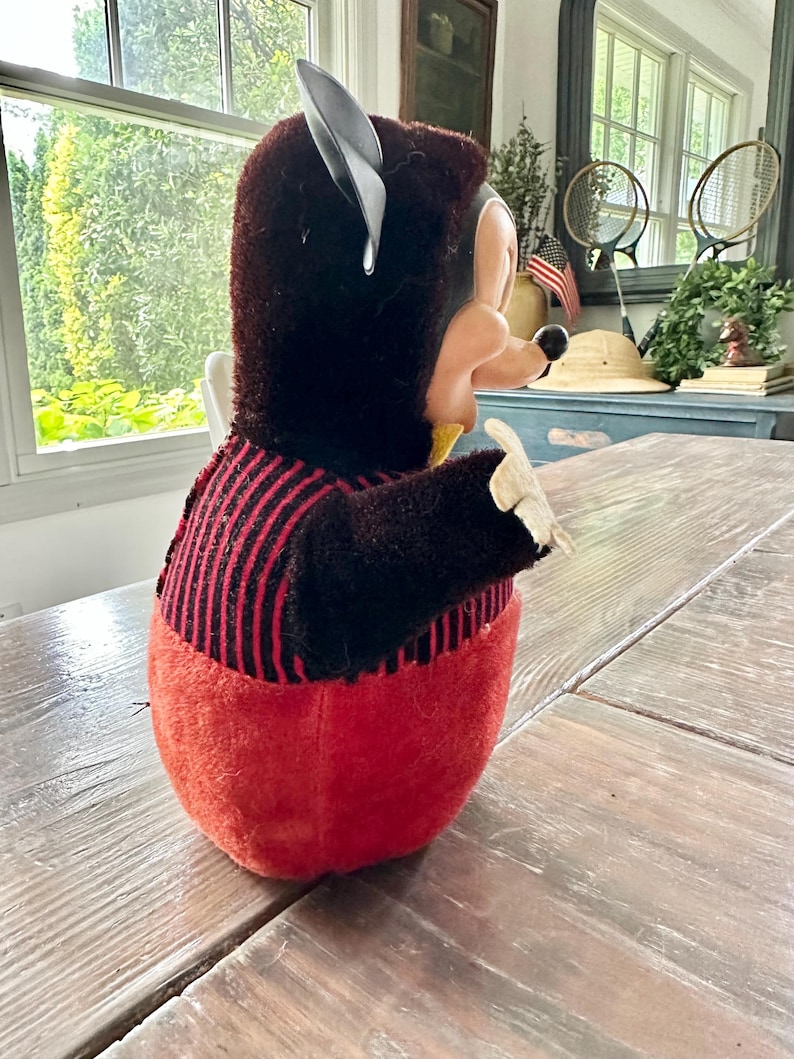 May include: A vintage plush toy in the shape of a cartoon character, featuring a black head with large ears and a red body. The toy has a black and red striped shirt and a yellow collar. The toy is sitting on a wooden table.
