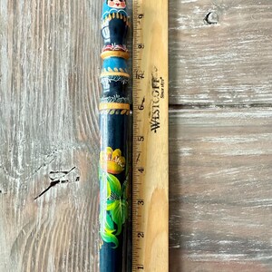May include: A hand-painted wooden pencil, approximately 9 inches long, featuring a Matryoshka doll design at the top. The pencil is dark blue with floral and geometric patterns in yellow, green, and red. A wooden ruler is next to the pencil.