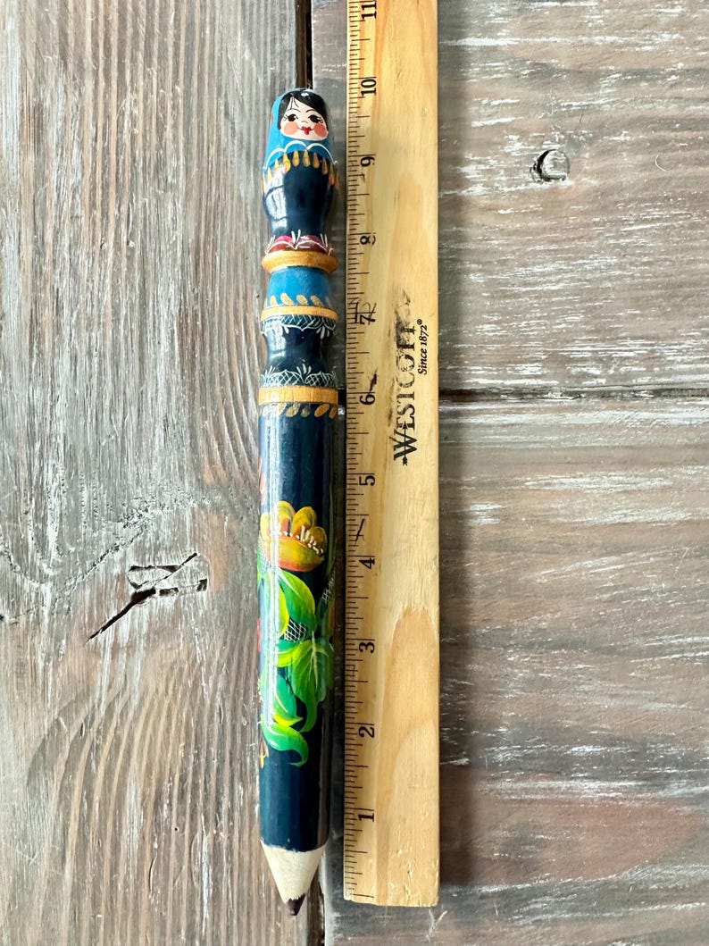 May include: A hand-painted wooden pencil, approximately 9 inches long, featuring a Matryoshka doll design at the top. The pencil is dark blue with floral and geometric patterns in yellow, green, and red. A wooden ruler is next to the pencil.