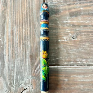 May include: A hand-painted pencil with a Russian nesting doll design on top. The pencil is dark blue with floral and geometric patterns in yellow, green, and gold. The pencil is resting on a weathered wooden surface.