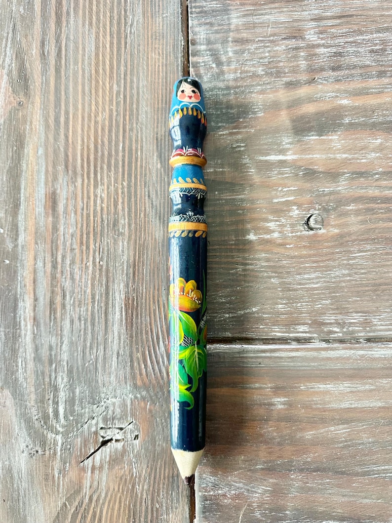 May include: A hand-painted pencil with a Russian nesting doll design on top. The pencil is dark blue with floral and geometric patterns in yellow, green, and gold. The pencil is resting on a weathered wooden surface.