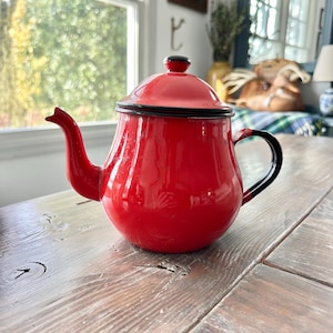 May include: A vibrant red enamel teapot with a black rim and handle. The teapot has a rounded body, a curved spout, and a lid with a black knob. It sits on a weathered wooden surface, suggesting a vintage or rustic aesthetic.
