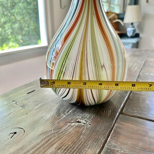 K&ouml;nnte beinhalten: Eine dekorative Glasvase mit Tropfenform. Die Vase hat vertikale Streifen in Blau-, Orange-, Gr&uuml;n- und Braunt&ouml;nen. Der Durchmesser des Vasenbodens betr&auml;gt ca. 15 cm.