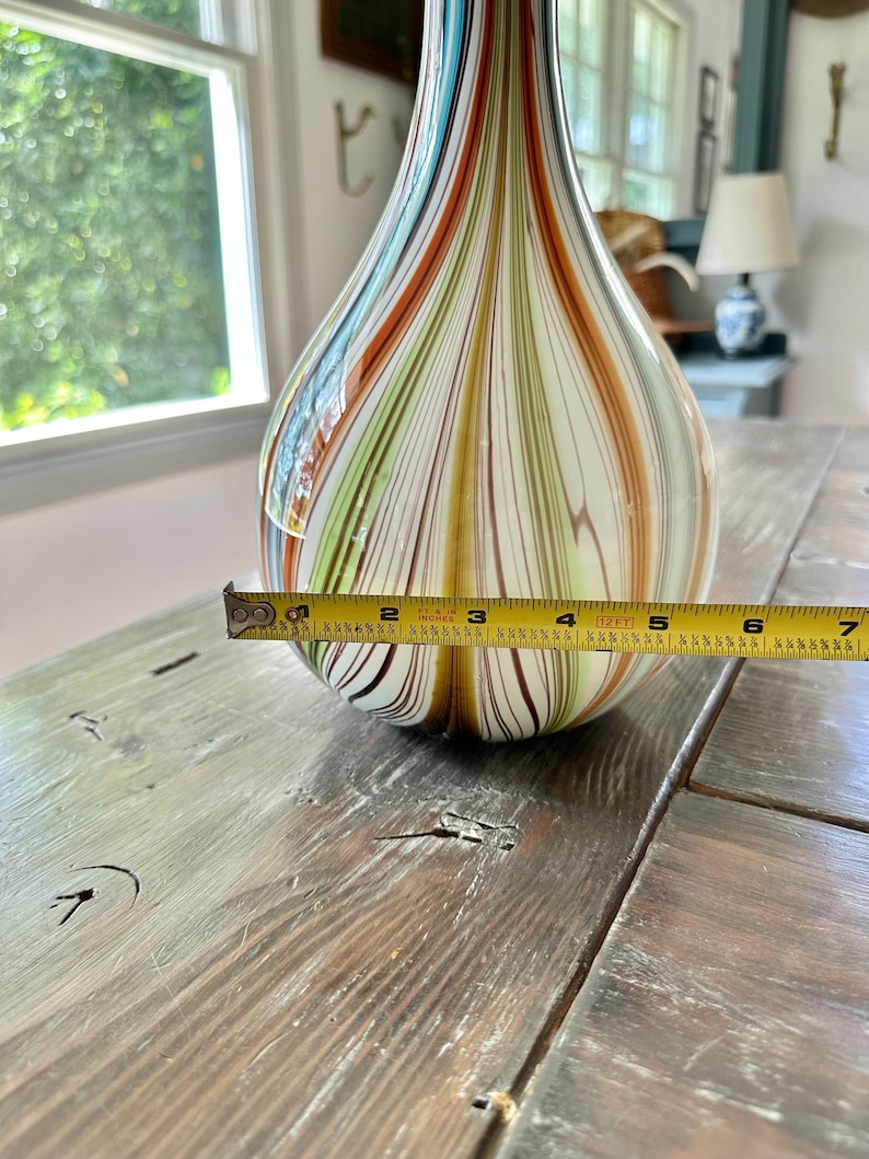 K&ouml;nnte beinhalten: Eine dekorative Glasvase mit Tropfenform. Die Vase hat vertikale Streifen in Blau-, Orange-, Gr&uuml;n- und Braunt&ouml;nen. Der Durchmesser des Vasenbodens betr&auml;gt ca. 15 cm.