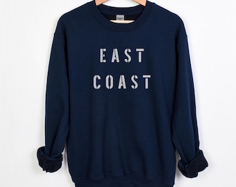 May include: A navy blue sweatshirt with the words "EAST COAST" printed in distressed white lettering. The sweatshirt is hanging on a wooden hanger. The sleeves are cuffed. The background is white.