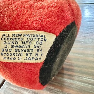 May include: Close-up of a vintage plush toy, primarily red with a black base. A tag is visible, with text indicating the toy's cotton contents and manufacturer, Gund Mfg. Co., made in Japan.