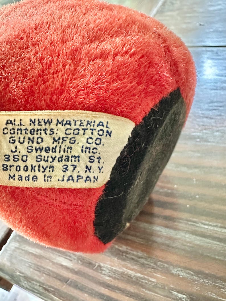 May include: Close-up of a vintage plush toy, primarily red with a black base. A tag is visible, with text indicating the toy's cotton contents and manufacturer, Gund Mfg. Co., made in Japan.