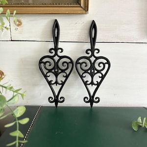 May include: Two black, heart-shaped trivets with ornate scrollwork and a handle. The trivets are made of cast iron and are displayed against a white wooden background. The trivets are approximately 7 inches long.