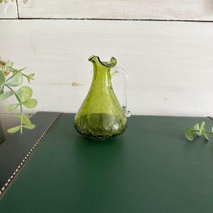May include: A small, green glass pitcher with a crackle finish. The pitcher has a handle and a ruffled rim. The pitcher is sitting on a dark green surface. Decorative item.
