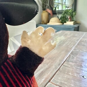 May include: A vintage plush toy, featuring a black hat and a red and black striped outfit. The toy's hand is visible, with a light-colored, worn fabric. The background includes a wooden table and a glimpse of a room with decor.