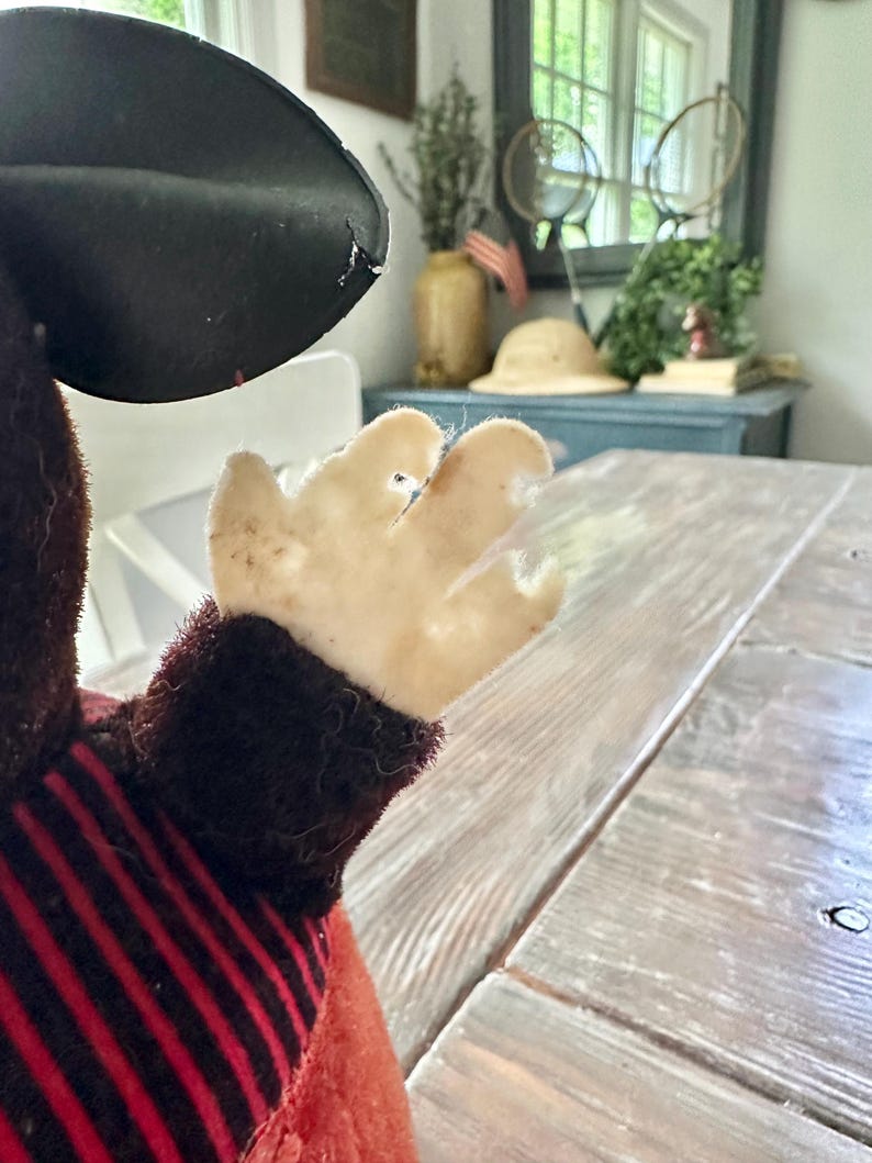 May include: A vintage plush toy, featuring a black hat and a red and black striped outfit. The toy's hand is visible, with a light-colored, worn fabric. The background includes a wooden table and a glimpse of a room with decor.