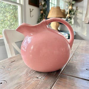 May include: A pink ceramic pitcher with a rounded body and a curved handle. The pitcher has a spout for pouring and sits on a weathered wooden table. The pitcher is a soft, muted pink color.