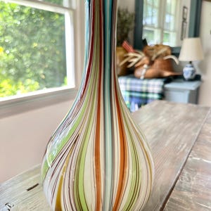 K&ouml;nnte beinhalten: Eine dekorative Glasvase mit tropfenf&ouml;rmiger Form und einem hohen, schlanken Hals. Die Vase hat vertikale Streifen in verschiedenen Farben, darunter Rot, Gr&uuml;n, Orange und Blau. Die Vase steht auf einer Holzoberfl&auml;che.
