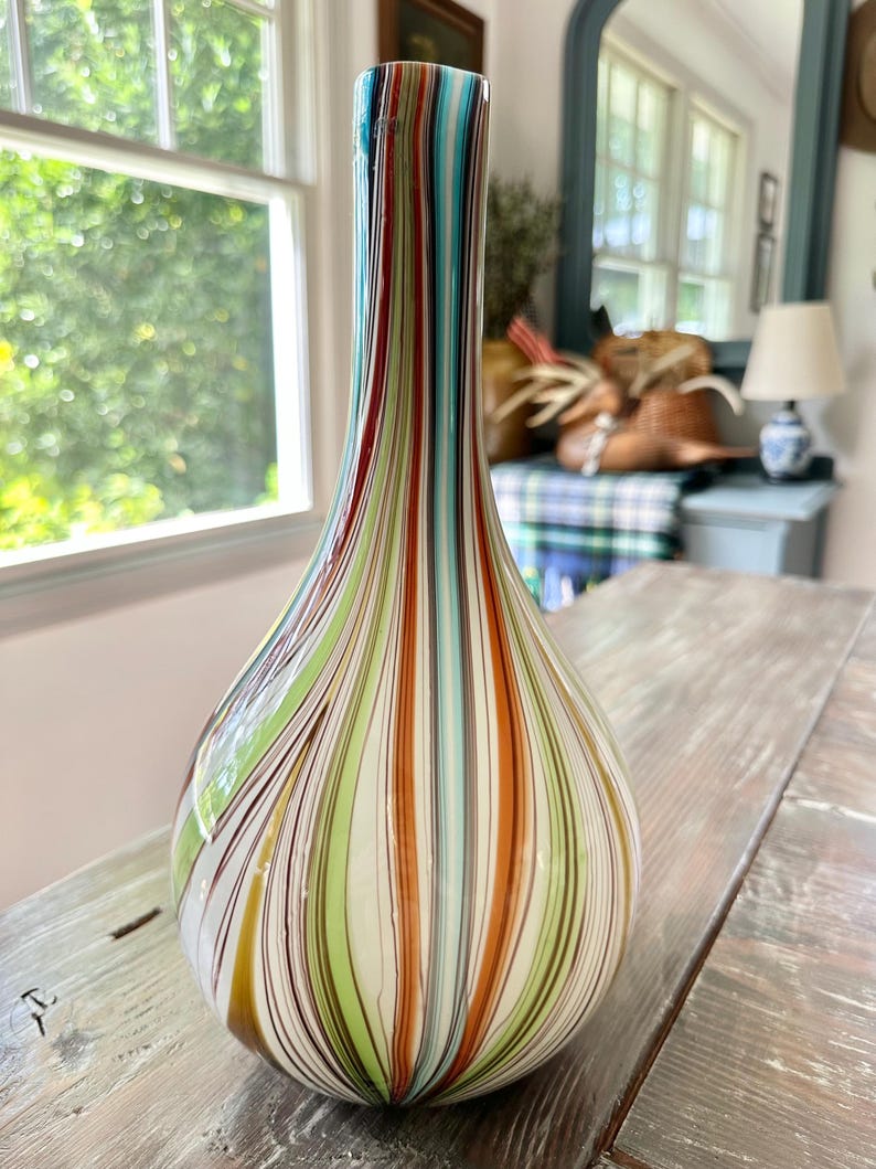 K&ouml;nnte beinhalten: Eine dekorative Glasvase mit tropfenf&ouml;rmiger Form und einem hohen, schlanken Hals. Die Vase hat vertikale Streifen in verschiedenen Farben, darunter Rot, Gr&uuml;n, Orange und Blau. Die Vase steht auf einer Holzoberfl&auml;che.