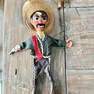 May include: A vintage marionette doll dressed in a traditional outfit, including a straw hat, dark green jacket, and gray pants with white trim. The doll holds a small silver toy gun. The marionette is suspended by strings attached to a wooden cross.