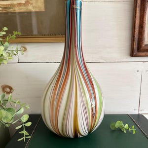 K&ouml;nnte beinhalten: Eine Murano-Glasvase mit bauchigem Boden und schlankem Hals. Die Vase hat vertikale Streifen in Blau-, Rot-, Orange-, Gr&uuml;n- und Braunt&ouml;nen. Die Vase ist etwa 30 cm hoch und steht auf einer dunkelgr&uuml;nen Oberfl&auml;che.