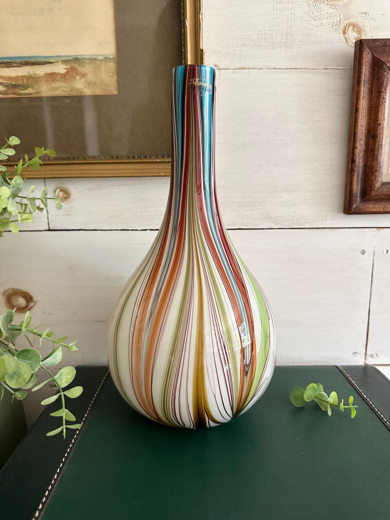 K&ouml;nnte beinhalten: Eine Murano-Glasvase mit bauchigem Boden und schlankem Hals. Die Vase hat vertikale Streifen in Blau-, Rot-, Orange-, Gr&uuml;n- und Braunt&ouml;nen. Die Vase ist etwa 30 cm hoch und steht auf einer dunkelgr&uuml;nen Oberfl&auml;che.