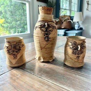 May include: Three handmade ceramic pieces with a rustic, earthy aesthetic. The set includes a tall, lidded bottle and two smaller vessels, all featuring expressive, carved faces. The pieces are in shades of tan and brown, with a textured finish.