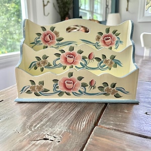 May include: A three-tiered wooden desk organizer painted cream with pink roses, green leaves, and blue ribbon accents. The organizer has a handle at the top and is designed to hold letters or documents. The edges are trimmed in blue.