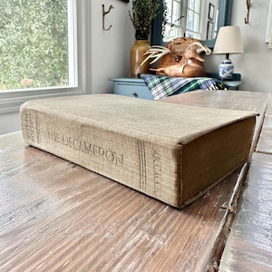 May include: A vintage book titled "The Decameron" with a tan linen cover, resting on a rustic wooden table. The book's spine features vertical lines and the publisher's name. The table has a weathered, distressed appearance.