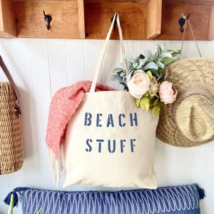 May include: Cream-colored tote bag with the words "BEACH STUFF" in navy blue. The bag is filled with a pink blanket, flowers, and is hanging on a wall with a straw hat and a woven bag.