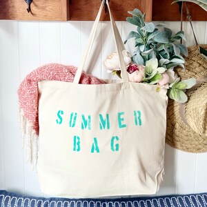 Puede incluir: Una bolsa de lona blanca con las palabras "SUMMER BAG" impresas en azul turquesa. La bolsa est&aacute; colgada de una estanter&iacute;a de madera con ganchos.
