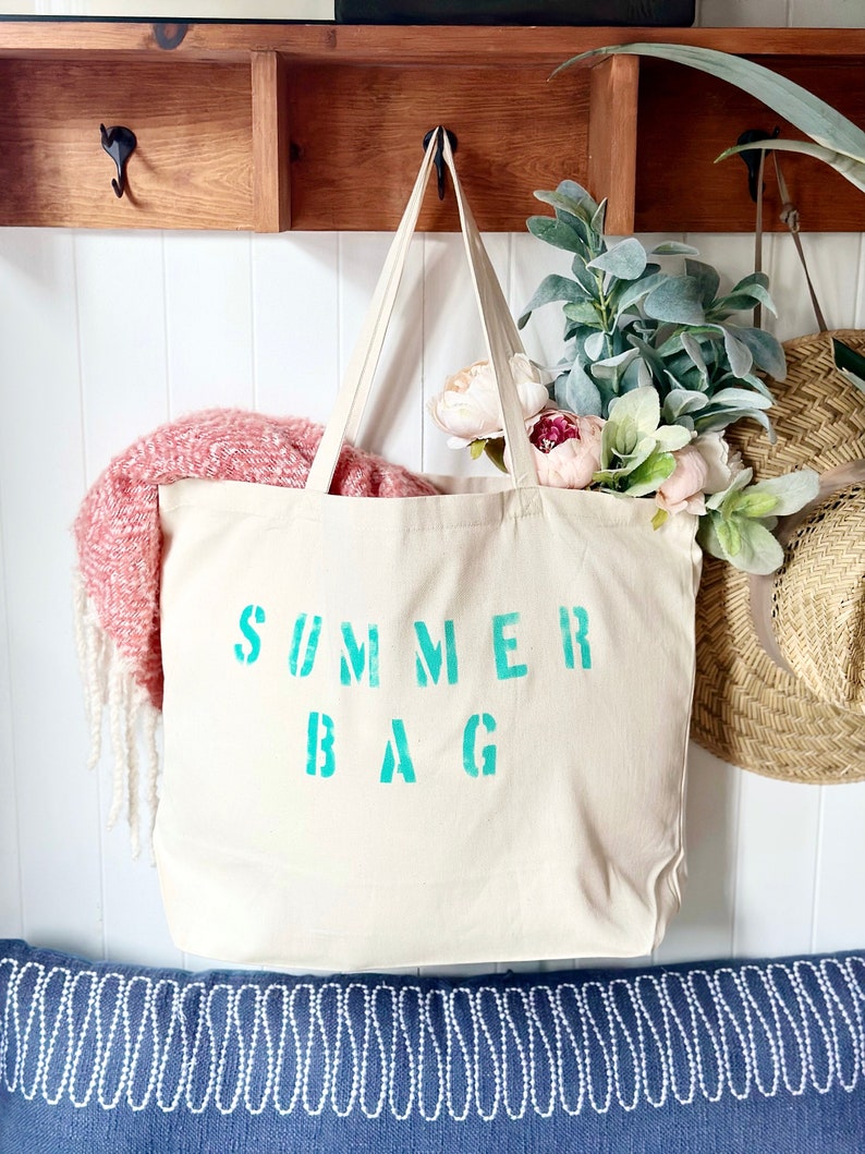 Puede incluir: Una bolsa de lona blanca con las palabras "SUMMER BAG" impresas en azul turquesa. La bolsa est&aacute; colgada de una estanter&iacute;a de madera con ganchos.