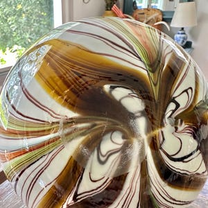 K&ouml;nnte beinhalten: Eine gro&szlig;e, runde Glasvase mit einem einzigartigen Wirbeldesign. Die Vase weist eine Kombination aus braunen, wei&szlig;en und schwarzen Streifen auf, die einen Marmoreffekt erzeugen. Die Farben verschmelzen und verleihen der Vase ein nat&uuml;rliches, organisches Aussehen. Die Vase ist ein dekorativer Gegenstand.