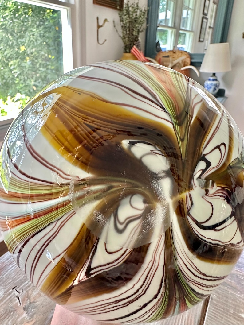 K&ouml;nnte beinhalten: Eine gro&szlig;e, runde Glasvase mit einem einzigartigen Wirbeldesign. Die Vase weist eine Kombination aus braunen, wei&szlig;en und schwarzen Streifen auf, die einen Marmoreffekt erzeugen. Die Farben verschmelzen und verleihen der Vase ein nat&uuml;rliches, organisches Aussehen. Die Vase ist ein dekorativer Gegenstand.