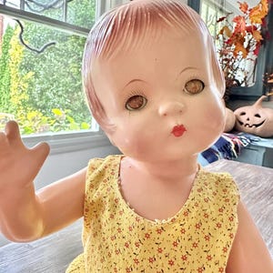 May include: A vintage baby doll with a painted face, brown eyes, and red lips. The doll wears a yellow dress with a floral pattern. The doll's arm is raised, and the background includes a window and decorative elements.
