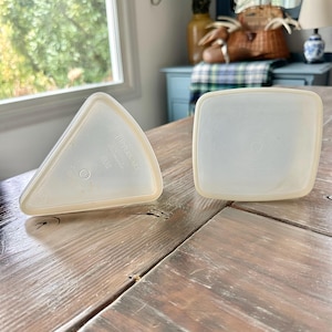 May include: Two vintage Tupperware containers on a wooden table. One is triangular, the other square, both with off-white lids and clear bases. The containers are empty and appear to be in good condition, with a light beige trim.
