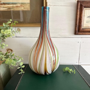 K&ouml;nnte beinhalten: Eine farbenfrohe Glasvase mit vertikalen Streifen in Blau-, Rot-, Orange-, Gr&uuml;n- und Wei&szlig;t&ouml;nen. Die Vase hat einen bauchigen Boden und einen schlanken Hals. Die Vase steht auf einer dunkelgr&uuml;nen Oberfl&auml;che.