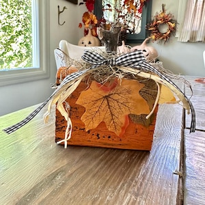 Könnte beinhalten: Eine orangefarbene Holzkiste, verziert mit Herbstblättern, getrocknetem Gras und einer schwarz-weiß karierten Schleife. Die Kiste ist ein Mittelstück auf einem Holztisch, mit einem Fenster und einem Spiegel im Hintergrund.