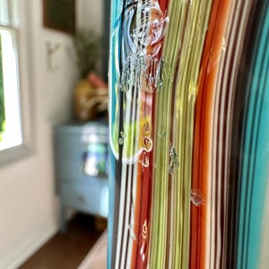 K&ouml;nnte beinhalten: Eine farbenfrohe Glasvase mit vertikalen Streifen in Rot-, Orange-, Gr&uuml;n-, Blau- und Braunt&ouml;nen. Die Vase hat eine strukturierte Oberfl&auml;che und ein klares, gl&auml;nzendes Finish. Der Hintergrund ist verschwommen und zeigt einen Blick in einen Raum.