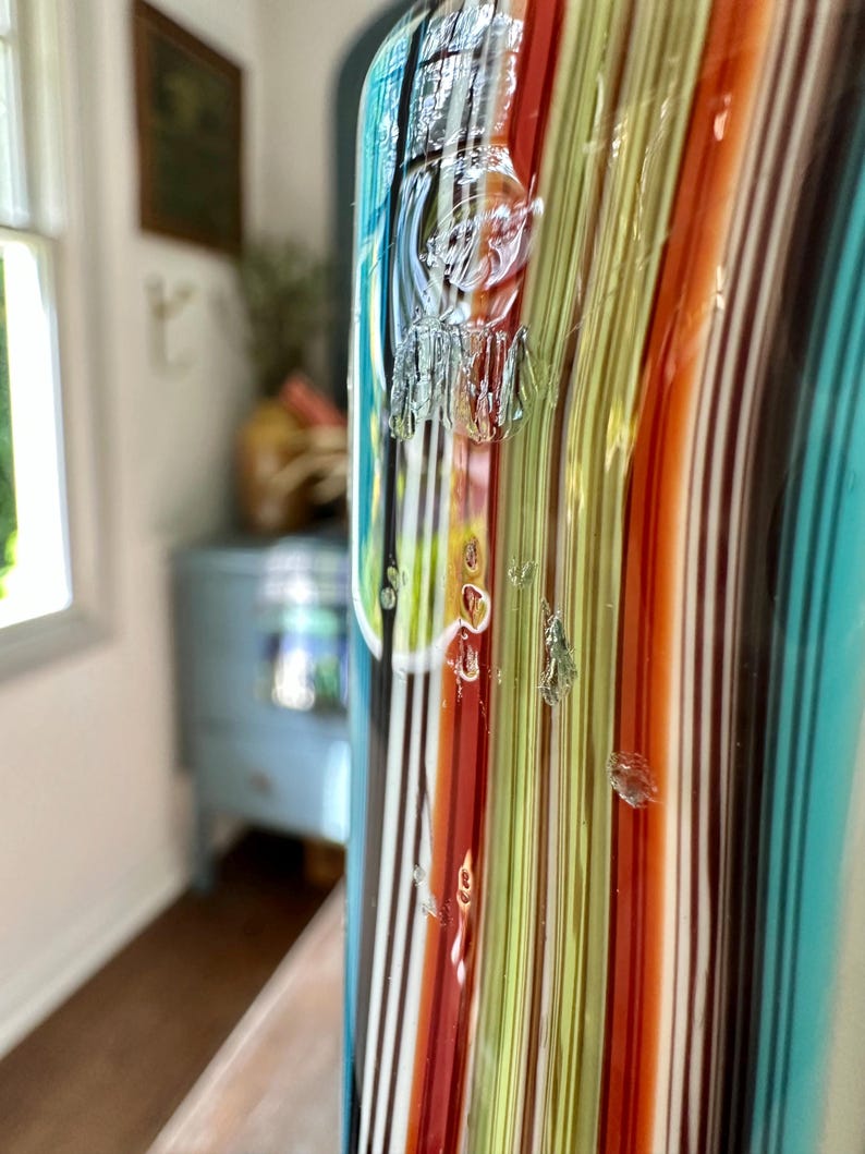 K&ouml;nnte beinhalten: Eine farbenfrohe Glasvase mit vertikalen Streifen in Rot-, Orange-, Gr&uuml;n-, Blau- und Braunt&ouml;nen. Die Vase hat eine strukturierte Oberfl&auml;che und ein klares, gl&auml;nzendes Finish. Der Hintergrund ist verschwommen und zeigt einen Blick in einen Raum.