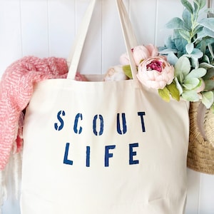 May include: A white canvas tote bag with blue lettering that reads "SCOUT LIFE". The bag is hanging on a wooden hook and has a pink blanket and flowers inside.