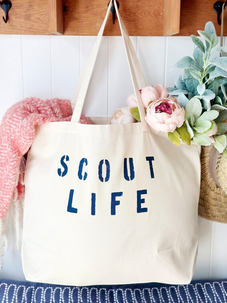 May include: A white canvas tote bag with blue lettering that reads "SCOUT LIFE". The bag is hanging on a wooden hook and has a pink blanket and flowers inside.
