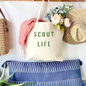 May include: A white canvas tote bag with the words "SCOUT LIFE" printed in green on the front. The bag is hanging on a wooden shelf with a pink scarf and a straw hat.