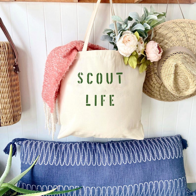 May include: A white canvas tote bag with the words "SCOUT LIFE" printed in green on the front. The bag is hanging on a wooden shelf with a pink scarf and a straw hat.