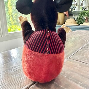May include: A vintage plush toy of a cartoon mouse, viewed from the back. The toy has large black ears, a brown head and arms, a red and black striped torso, and red pants. The toy is sitting on a wooden table.