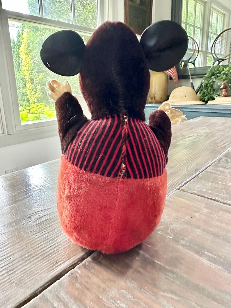 May include: A vintage plush toy of a cartoon mouse, viewed from the back. The toy has large black ears, a brown head and arms, a red and black striped torso, and red pants. The toy is sitting on a wooden table.